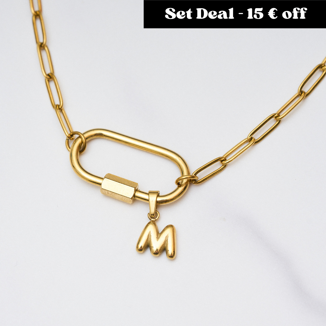 Goldene Gliederkette mit großem Karabinerverschluss, an dem ein herzförmiger Anhänger mit Perlenrand und ein kleiner Buchstabe „M“ hängen. Oben rechts steht in einem schwarzen Kasten: „Set Deal – 15 € off