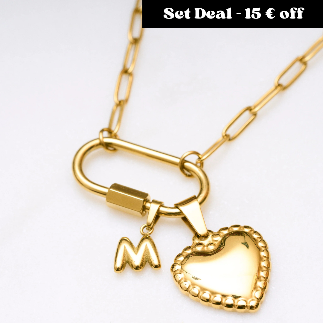 Goldene Gliederkette mit großem Karabinerverschluss, an dem ein herzförmiger Anhänger mit Perlenrand und ein kleiner Buchstabe „M“ hängen. Oben rechts steht in einem schwarzen Kasten: „Set Deal – 15 € off